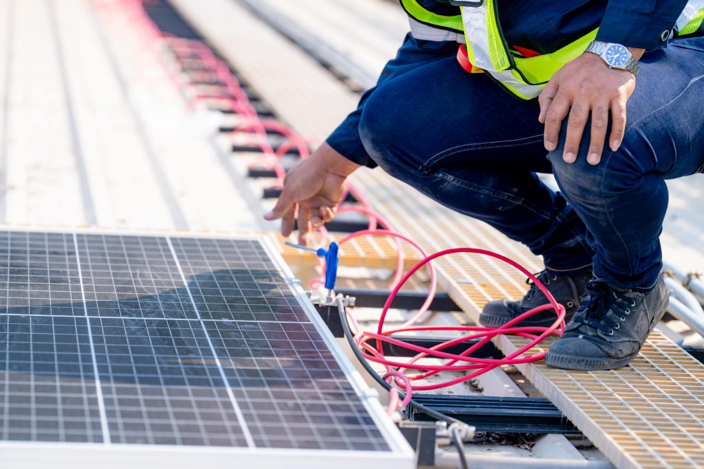 Eine Person in Schutzausrüstung kniet auf einem Dach und verbindet während der Installation rote Solarleitungsdrähte mit einem Solarpanel.