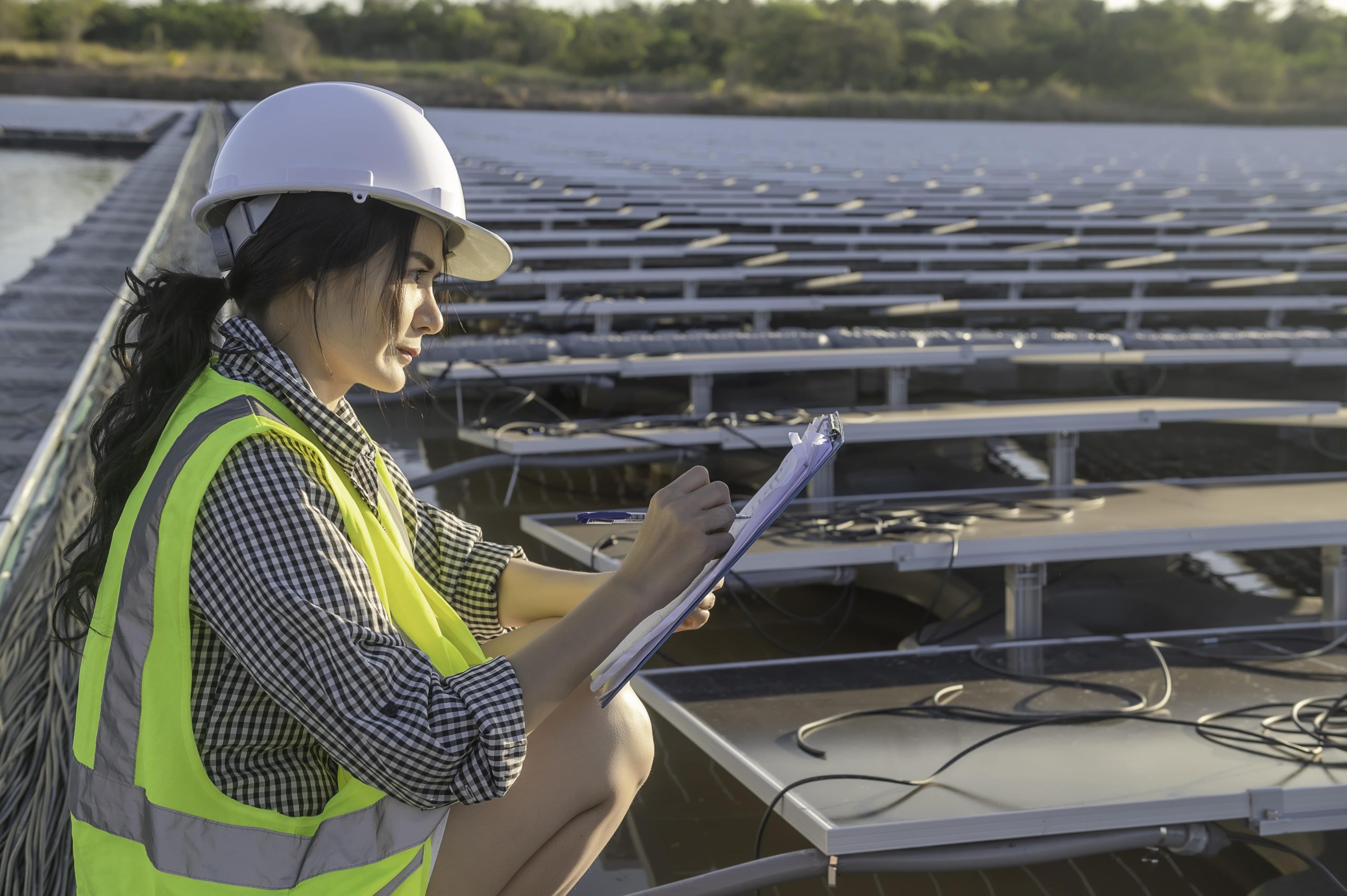 Eine Person mit Schutzhelm und Sicherheitsweste schreibt auf einem Klemmbrett, während sie Solarmodule einer Außenanlage inspiziert.