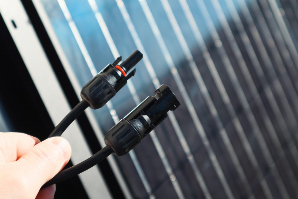 Eine Hand hält zwei Solarpanel-Anschlüsse vor Photovoltaik-Solarpanelzellen.