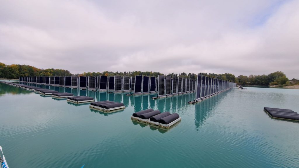 Auf einem ruhigen See, umgeben von Bäumen und unter einem wolkenverhangenen Himmel, sind Reihen von schwimmenden Solarzellen installiert.
