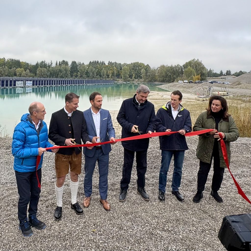 Sechs Personen stehen im Freien auf Kies an einem See und schneiden bei einer Eröffnungsfeier ein rotes Band mit einer Schere durch. Im Hintergrund sind Bäume und eine schwimmende PV-Anlage zu sehen.