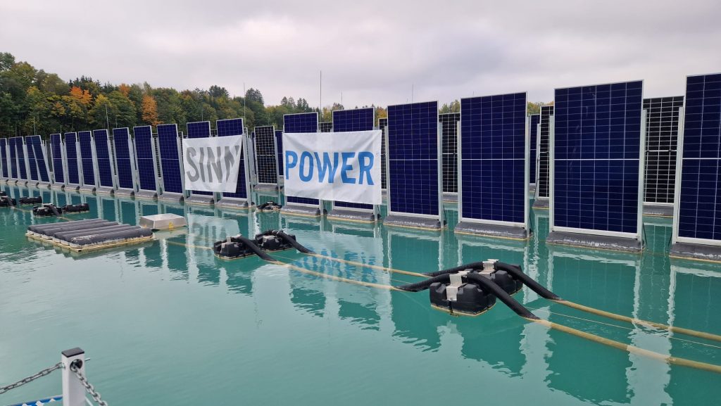 Reihen von schwimmenden Solarpaneelen auf einem Gewässer, mit zwei großen Bannern, auf denen "SINN" und "POWER" steht. Bäume und bewölkter Himmel im Hintergrund.