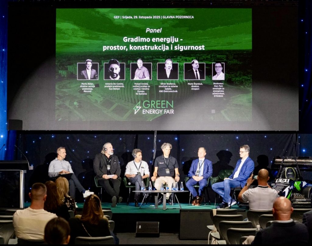 Sechs Podiumsteilnehmer sitzen auf der Bühne und diskutieren über Energie, Bauwesen und Sicherheit auf der Green Energy Fair, während hinter ihnen ein großes Veranstaltungsbanner zu sehen ist. Im Vordergrund ist das Publikum zu sehen.