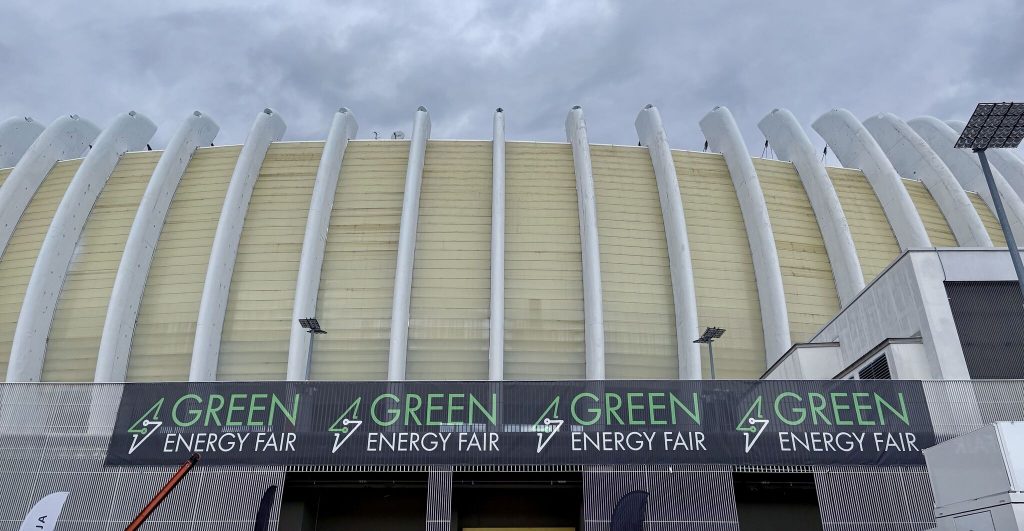 Großes Gebäude mit geschwungenen vertikalen Strukturen und einem Banner mit der Aufschrift "Green Energy Fair", das mehrfach über der Fassade angebracht ist, unter einem bewölkten Himmel.