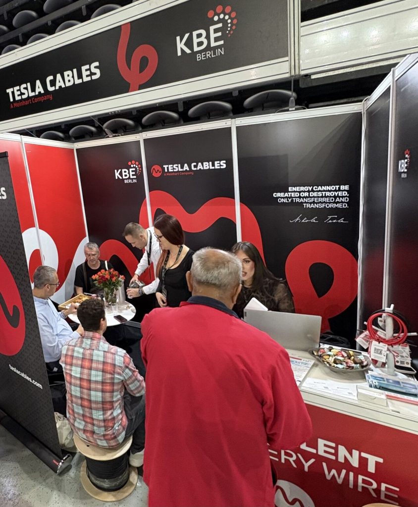 Eine Gruppe von Menschen unterhält sich und arbeitet an einem Messestand von Tesla Cables und KBE Berlin, wo Werbematerial und Kabel auf Tischen ausgestellt sind.