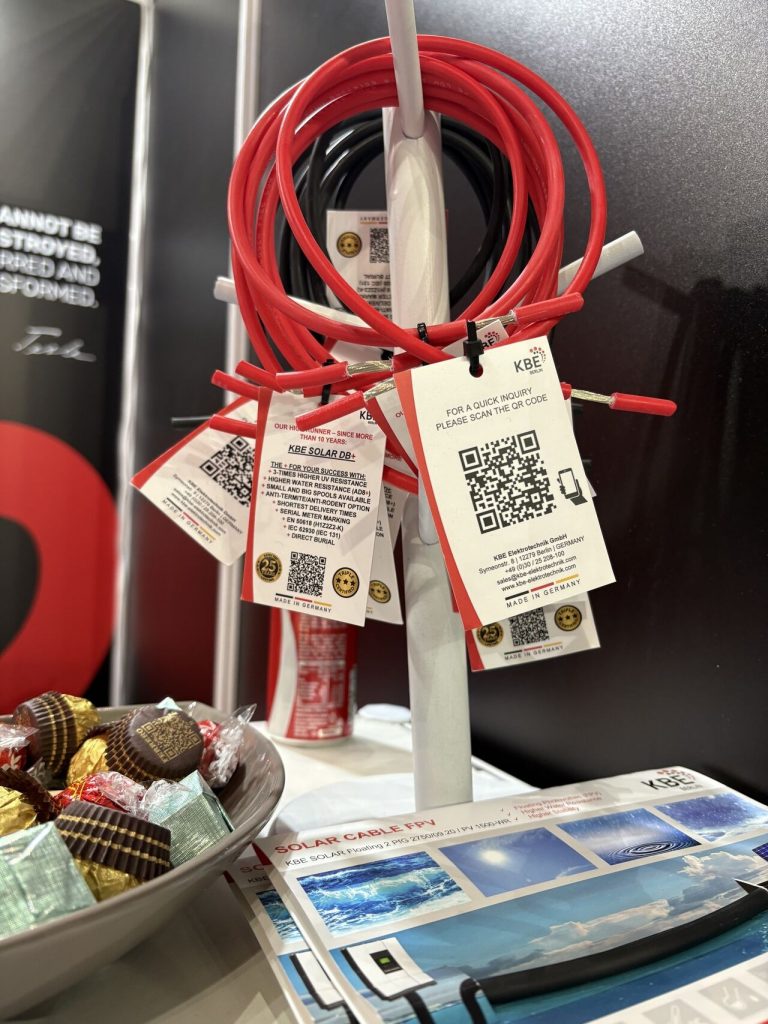 Ein Ständer mit roten und schwarzen Ladekabeln von KBE Berlin, die jeweils mit QR-Code-Tags versehen sind, neben einer Schale mit verschiedenen Pralinen und einigen Broschüren auf einem Tisch.