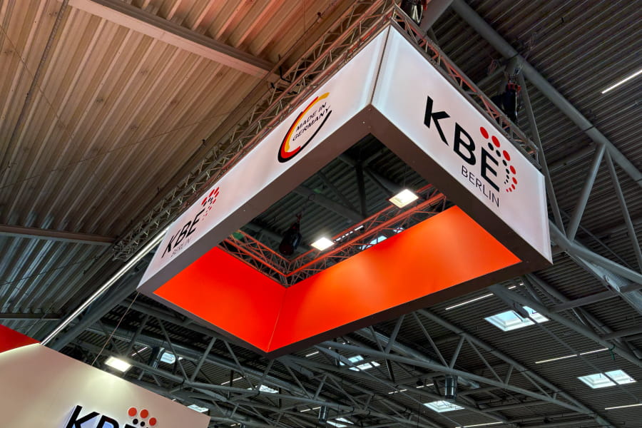 Eine große hängende Ausstellungskonstruktion mit dem KBE-Berlin-Logo und dem „German Innovation“-Branding, die an der Decke einer mit Metallträgern ausgestatteten Ausstellungshalle aufgehängt ist.